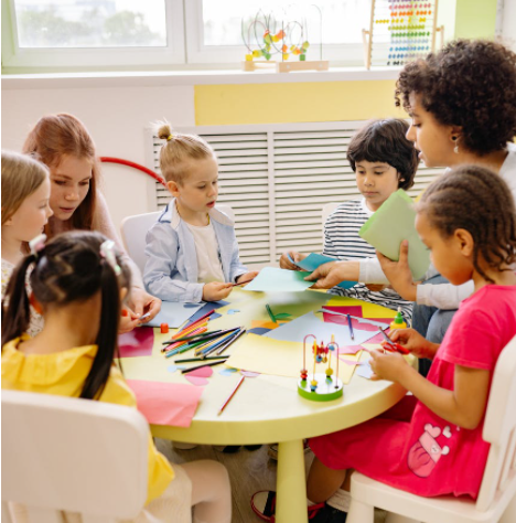 early childhood teachers and children sitting at a low table with paper and pencils.PNG